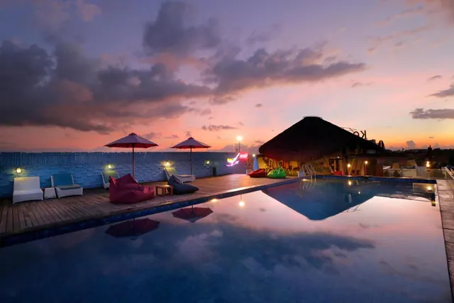 A rooftop swimming pool at dusk with red parasol umbrellas, white sun loungers, bean bags and a thatched-roof structure under a pink and purple sky.