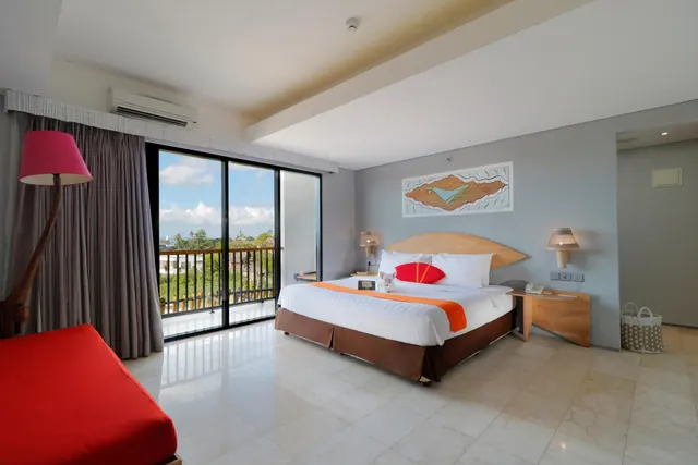 A bedroom with a king bed, a red sofa, a wall-mounted air conditioner, and sliding glass doors opening onto a balcony with a view of trees.