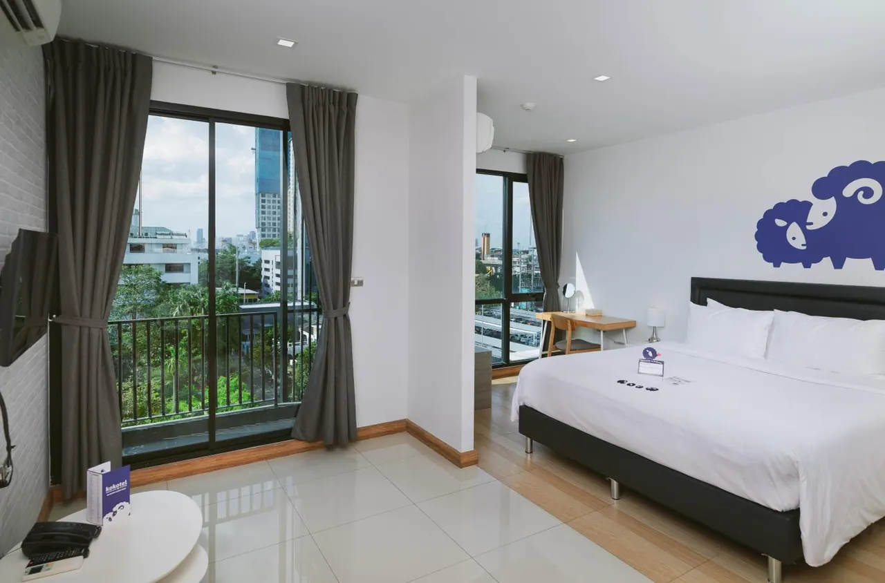 A bedroom with a large white bed, a sliding glass door opening onto a balcony with city buildings and trees beyond, a wooden desk, and a blue sheep wall decal.