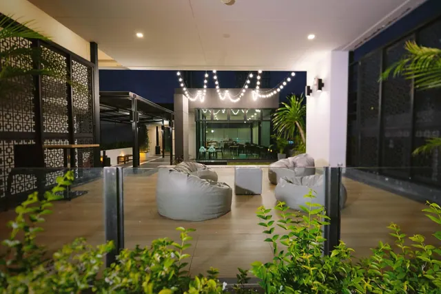 A covered outdoor terrace at night with bean bag seating, string lights and plants along a glass railing, with a lit indoor dining area visible beyond.