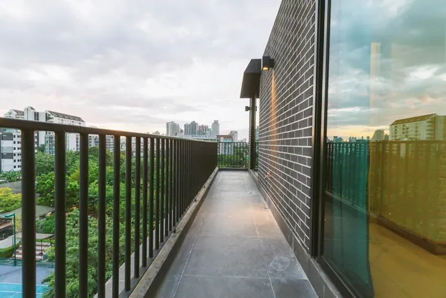 A narrow tiled balcony runs alongside a dark brick wall and large glass doors, with a black metal railing overlooking trees, low buildings and a city skyline under a cloudy sky.
