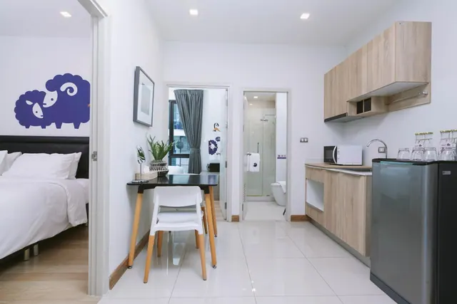 A small apartment-style room with a black dining table and white chair, a kitchenette with a microwave, sink and fridge, and open doorways into a bedroom and a bathroom with a glass-walled shower.