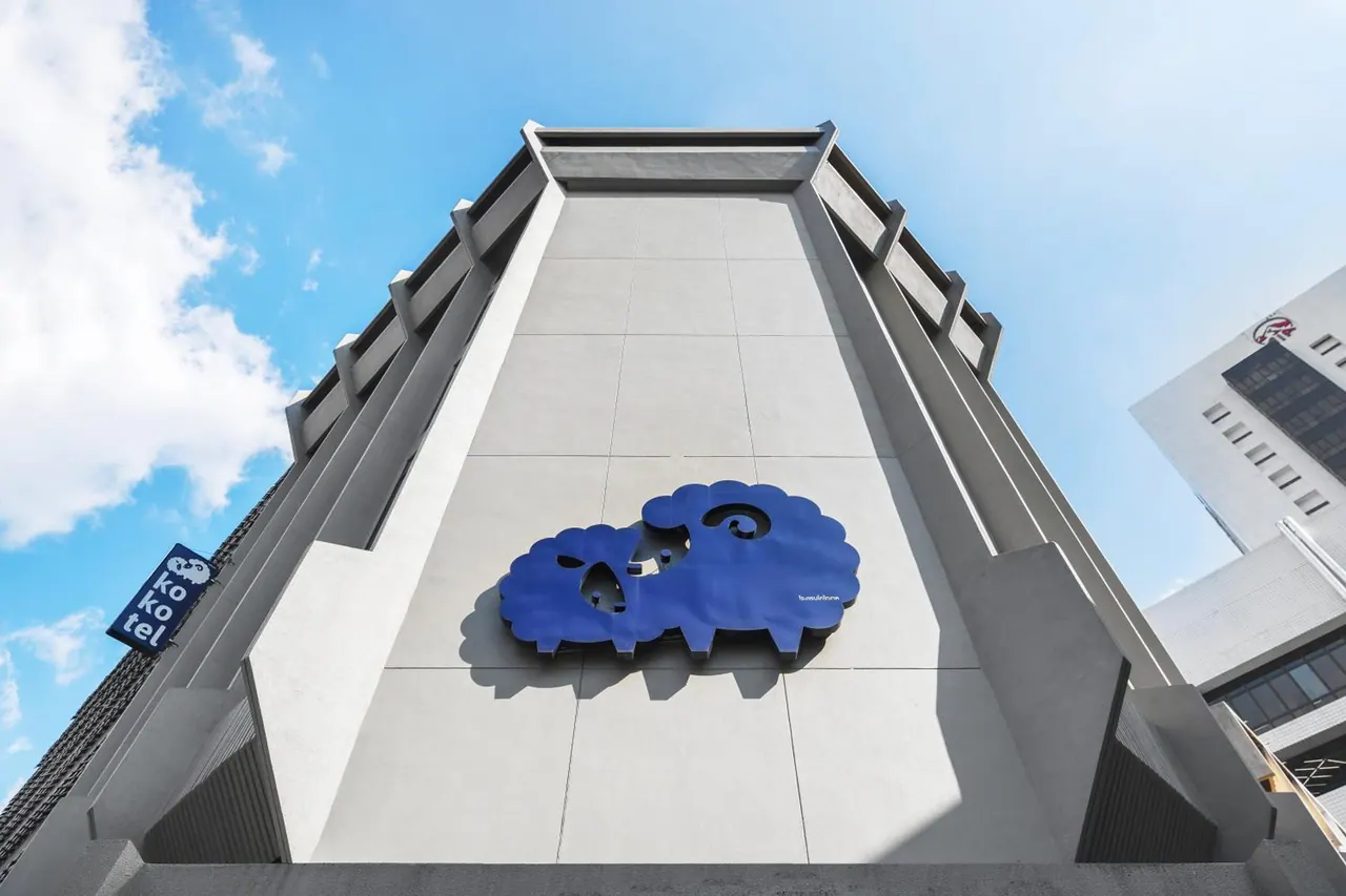 Low angle view of a white concrete building facade with a large blue sheep logo and a kokotel sign under a blue sky.