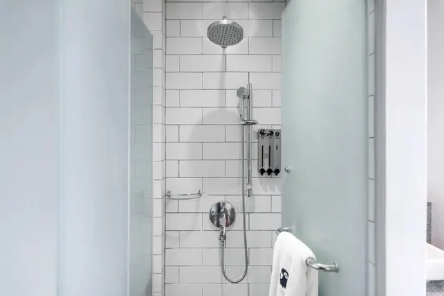 A walk-in shower with white subway tiles, a rain shower head and handheld shower, wall-mounted soap dispensers, and a towel hung on a grab bar beside a frosted glass door.