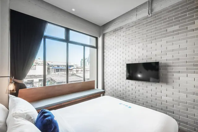 A bedroom with a made bed, a wall-mounted television on a textured brick-style wall, and a window looking out over low-rise city buildings.