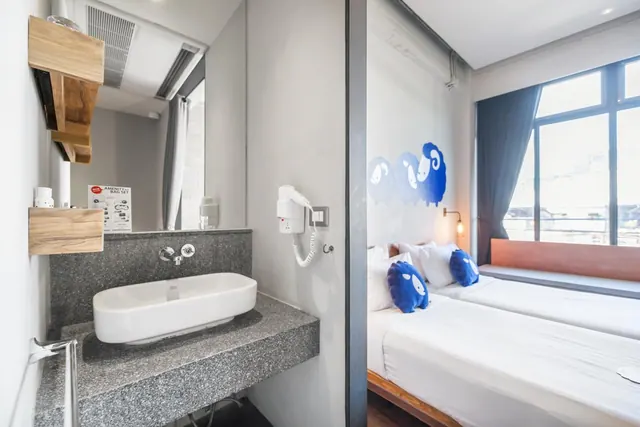 A bathroom with a vessel sink on a granite counter and a wall-mounted hair dryer, open to a bedroom with two beds, blue sheep pillows and a window with a bench seat.