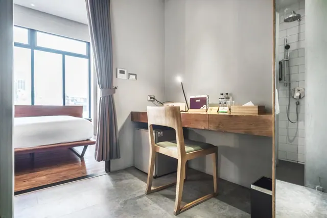 A bedroom with a wooden desk and chair, a small fridge beneath the desk, and a glass-walled walk-in shower visible on the right.