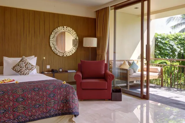 A bedroom with a made bed decorated with frangipani flowers, a red armchair, a round ornate mirror, and sliding glass doors opening onto a balcony with a daybed and tropical greenery.
