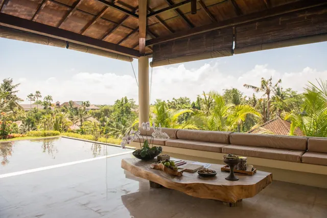 A covered terrace with a long built-in sofa and a live-edge wooden coffee table sits beside an infinity pool and a view of tropical trees.