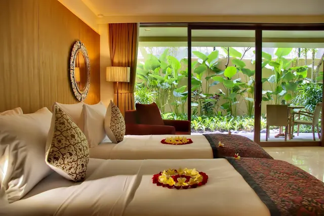 A bedroom with two beds decorated with flower arrangements, a seating armchair, and sliding glass doors opening onto a garden with an outdoor table and chairs.
