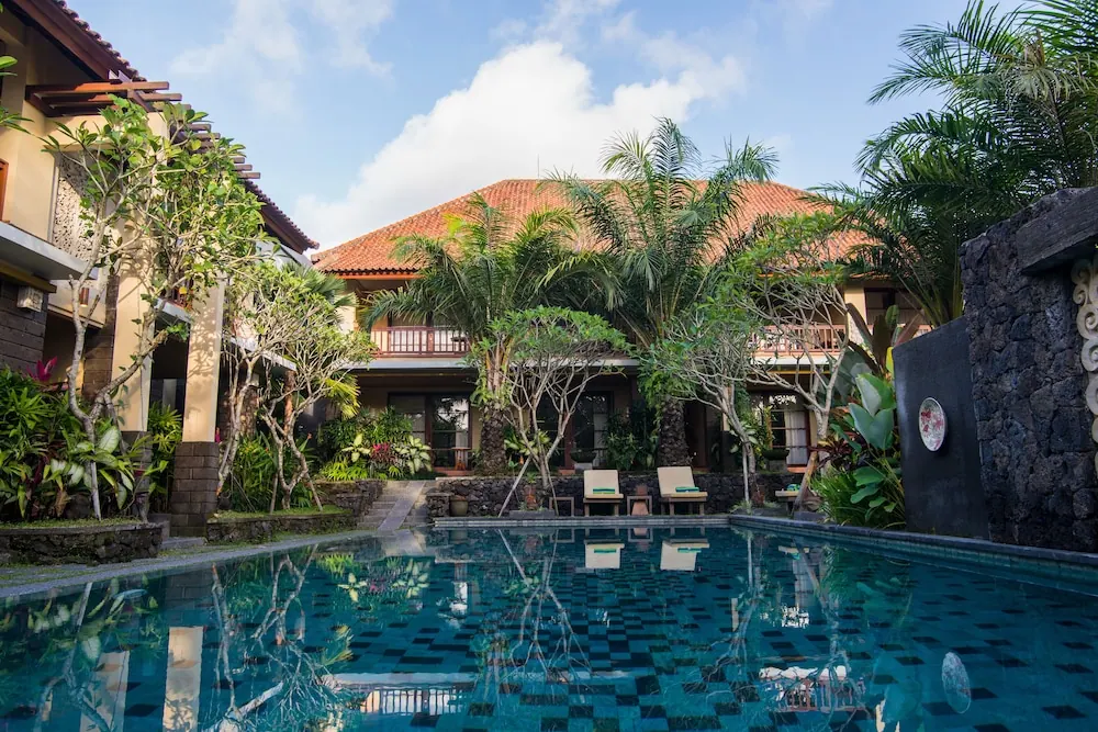 A tiled courtyard swimming pool with two white sun loungers, surrounded by palm and frangipani trees and a two-storey building with a terracotta roof.