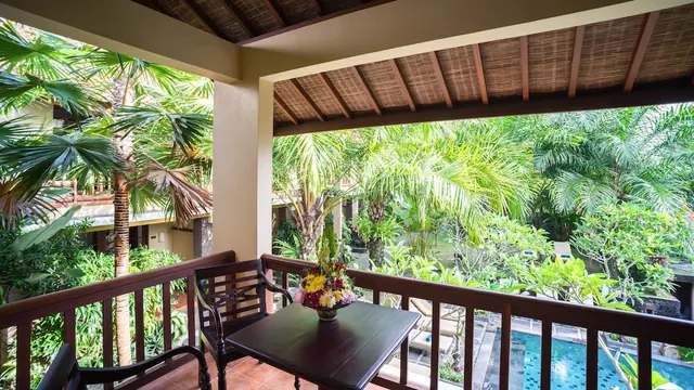 A covered balcony with a dark wooden table, chairs and a flower arrangement overlooks a tropical garden and a swimming pool with sun loungers.