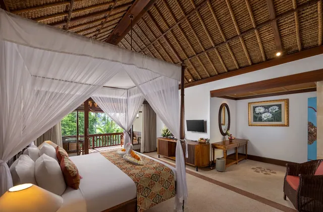 A bedroom with a four-poster bed under white netting, a thatched ceiling, a wall-mounted television, and a balcony with greenery visible through the glass doors.