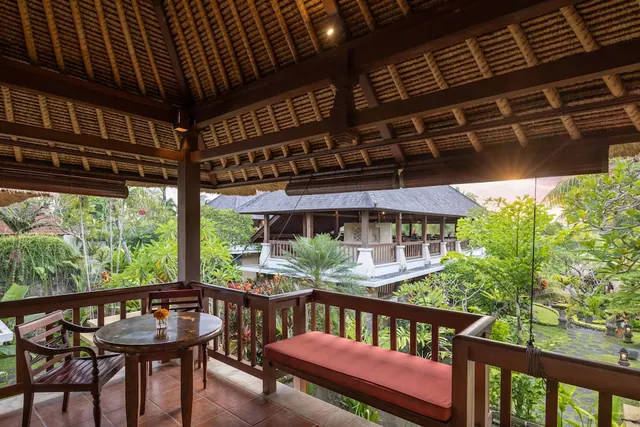 A covered terrace with a round table, two wooden chairs and a red-cushioned bench overlooks tropical gardens and a pavilion at sunset.