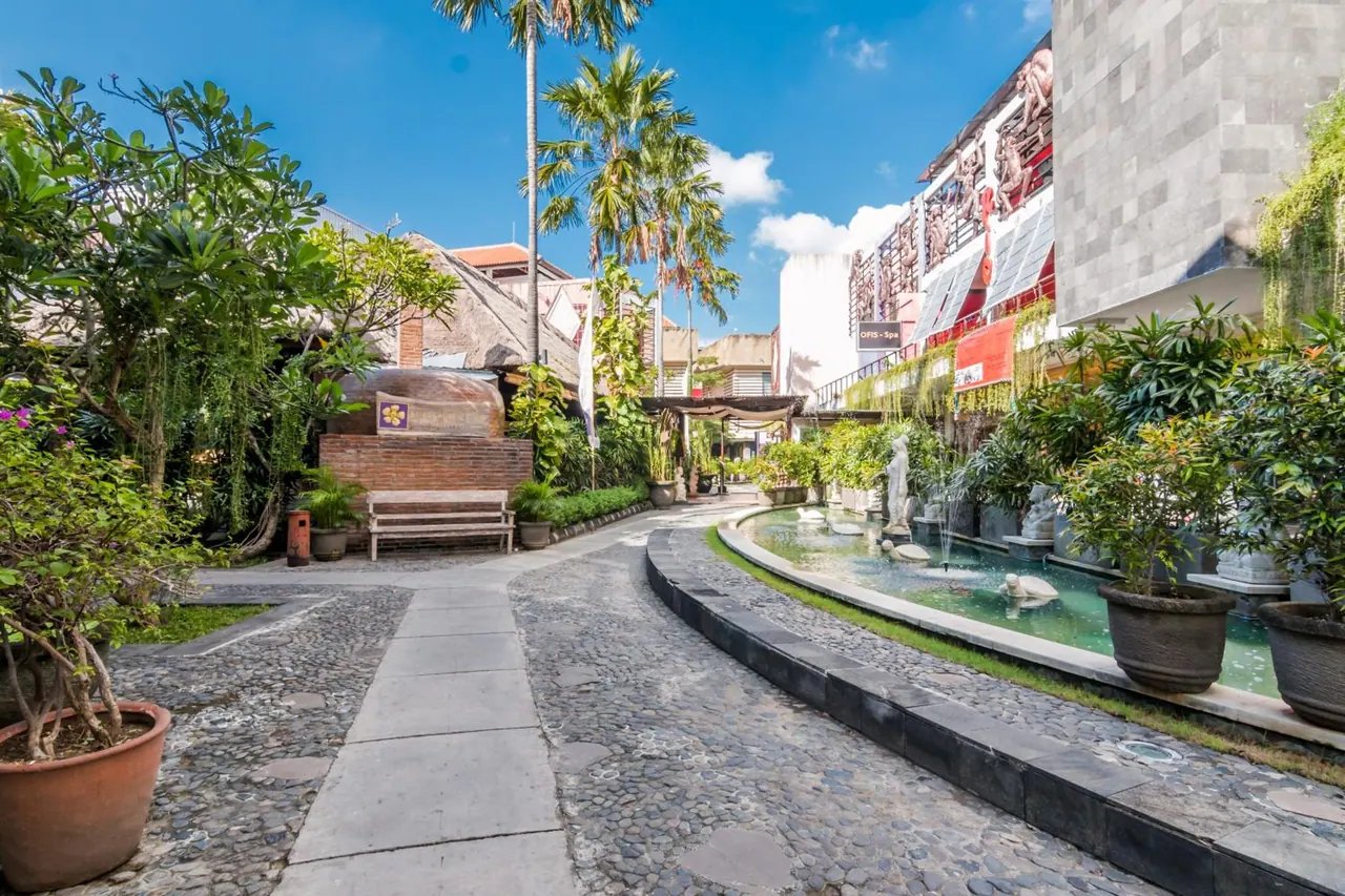 A paved garden walkway with a cobblestone path runs past a small fountain pool with statues, potted plants, palm trees and a wooden bench, beside a stone-tiled building with a spa sign.