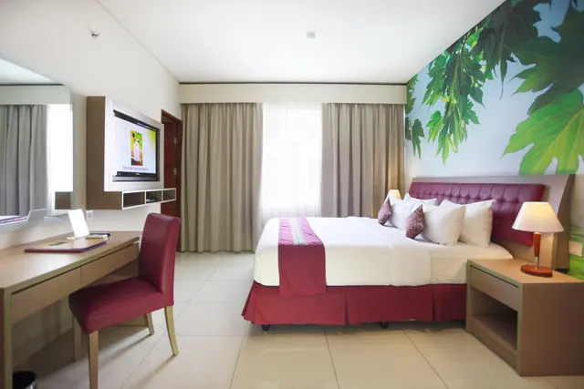 A hotel bedroom with a king bed, a leaf-patterned wall mural, a wall-mounted television and a desk with a red chair.