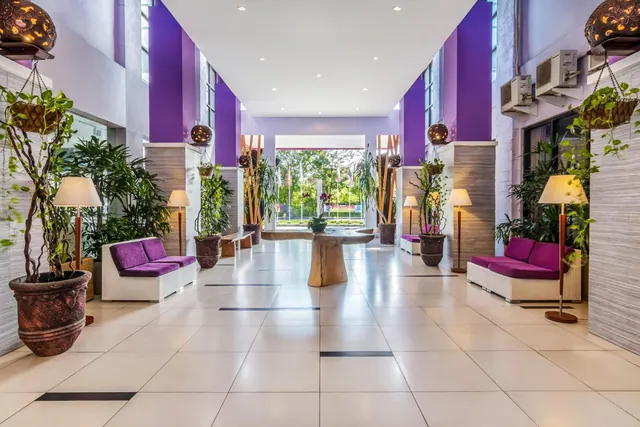 A symmetrical view down a bright lobby with purple walls, white wicker sofas, potted plants and a wooden table, with greenery visible through the glass at the far end.