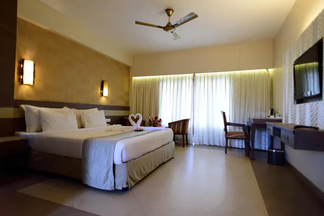 A spacious hotel bedroom contains a double bed, desk, seating, television, ceiling fan and bright curtained window.