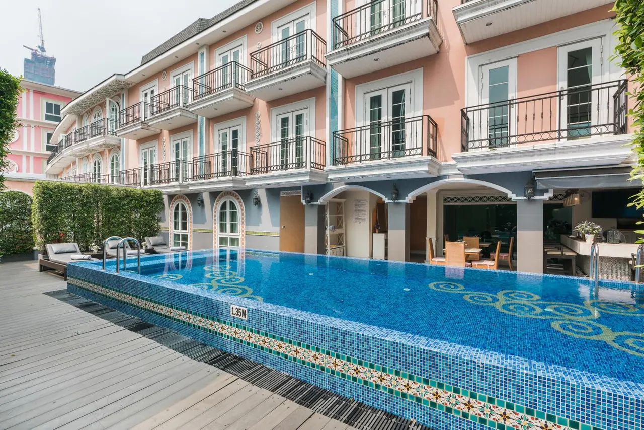 Outdoor swimming pool with blue mosaic tiles beside a pink multi-storey building with balconies, with sun loungers on the wooden deck and an open restaurant under arches at ground level.