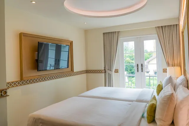 A bedroom with two white beds, a wall-mounted television, and French doors opening onto a balcony with trees and buildings beyond.