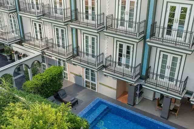 Elevated view of a white hotel building with balconies and French doors, with a blue pool and sun loungers below.