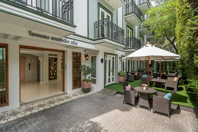 Exterior of a white hotel building with green-trimmed balconies, showing a lawn courtyard with outdoor tables, chairs and umbrellas beside a tiled entrance path.