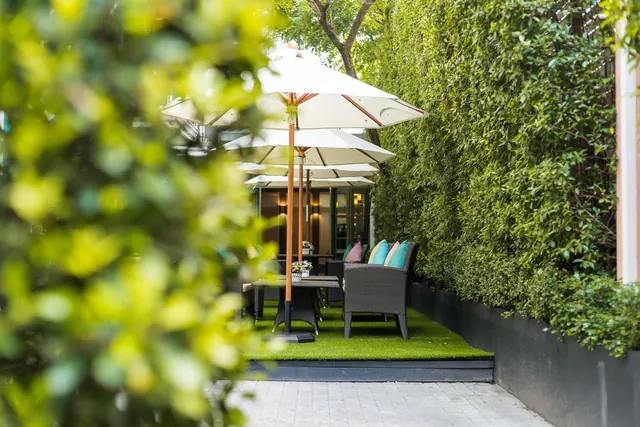 Outdoor seating area with white parasol umbrellas, a wicker sofa with teal cushions and a table on artificial grass beside a tall hedge.