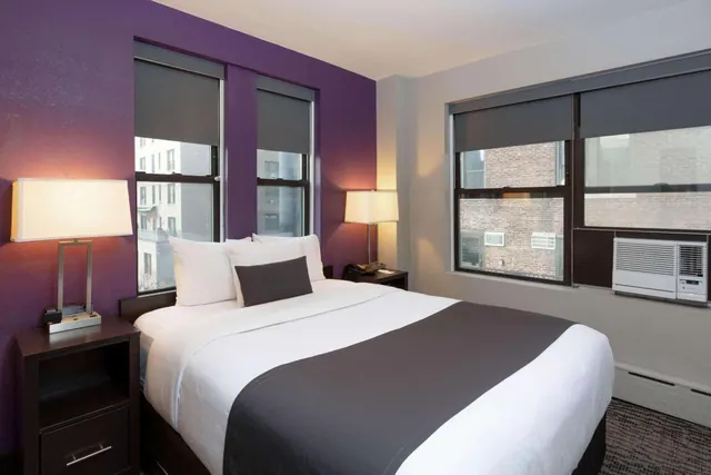 A bedroom with a double bed dressed in white linen and a grey runner, a purple accent wall, two bedside lamps, and windows above a window air conditioner, looking onto a brick building.