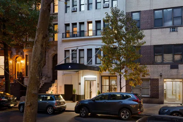 Street view of the hotel entrance at dusk, with a dark awning over the doors, parked cars along the curb, and a tree on the sidewalk.