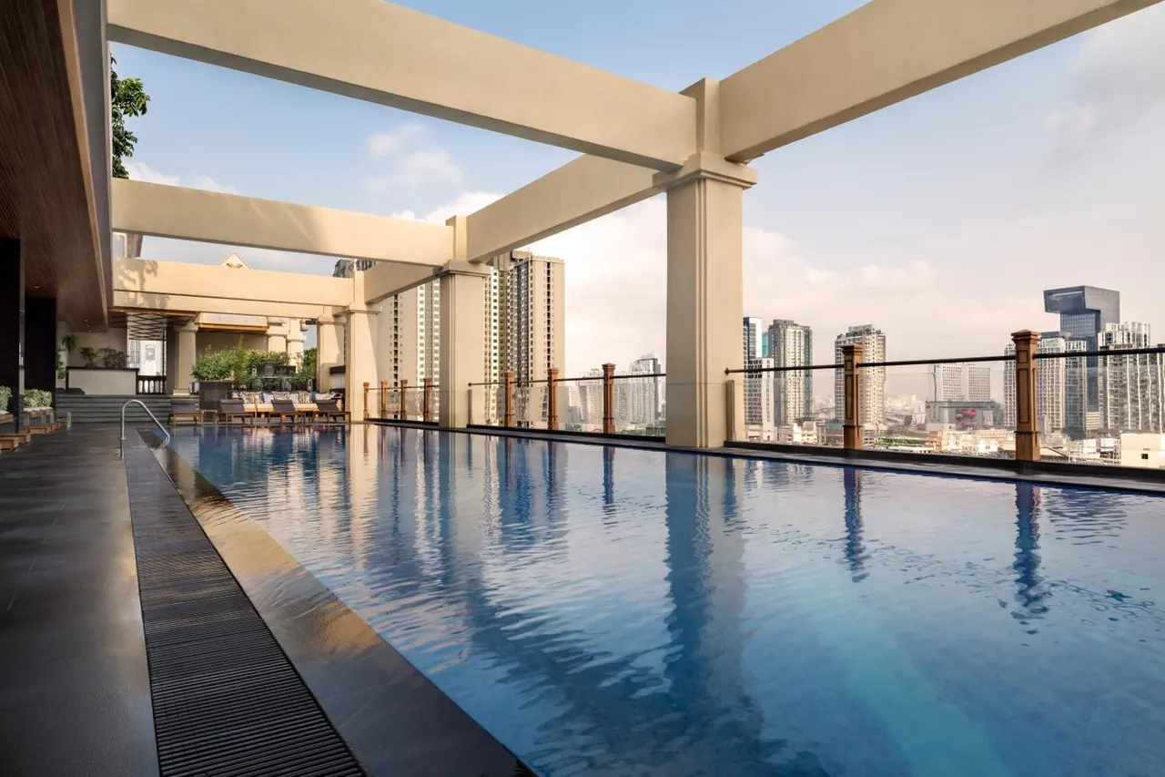 A long rooftop swimming pool with beige pergola beams, sun loungers in the background, and a view of high-rise buildings under a hazy sky.