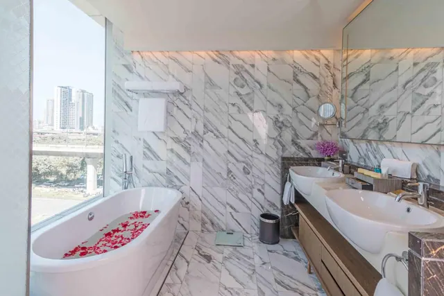 A marble bathroom with a freestanding bathtub scattered with red rose petals, a double vanity, and a large window overlooking city high-rises and an elevated road.