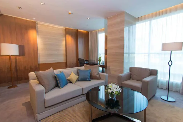 A living area with a grey sofa and armchair, a glass coffee table with white flowers, two floor lamps, a desk in the background, and sheer-curtained windows.