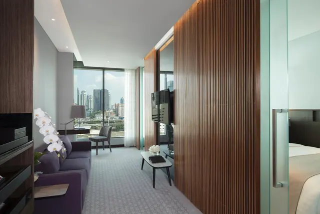 A hotel living area with a purple sofa, a white orchid, a wooden slatted wall, a wall-mounted television, and floor-to-ceiling windows overlooking city towers, with a bed visible through a glass door on the right.