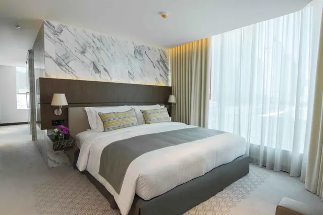 A bedroom with a king bed, a marble feature wall, and floor-to-ceiling windows with sheer curtains overlooking hazy city buildings.