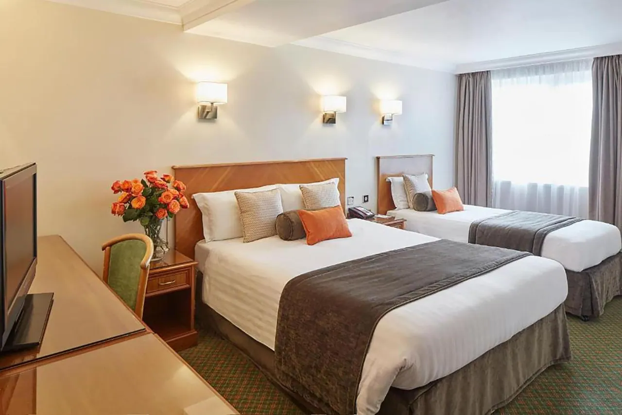 A hotel bedroom with a double bed and a single bed, wall lights, a flat-screen television, a green chair and a vase of orange roses.