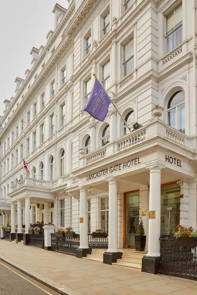 The white stucco facade of the Lancaster Gate Hotel is shown with a purple flag, a Union Jack, white columns, a balcony and black railings at street level.