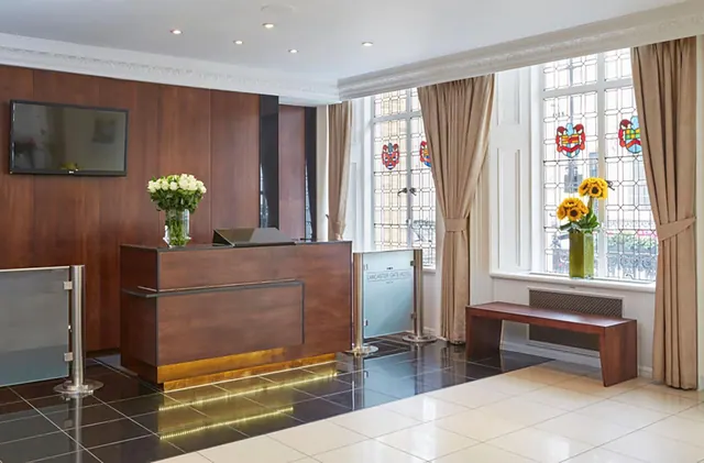 A hotel reception area with a wood-panelled desk, flower arrangements, a wall-mounted television, a wooden bench and stained glass windows.