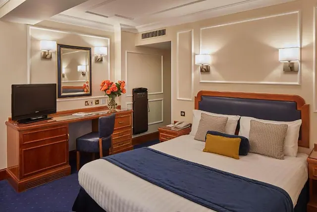 A double bed with a navy runner and cushions sits beside a wooden desk with a television, mirror and vase of orange roses in a hotel bedroom.