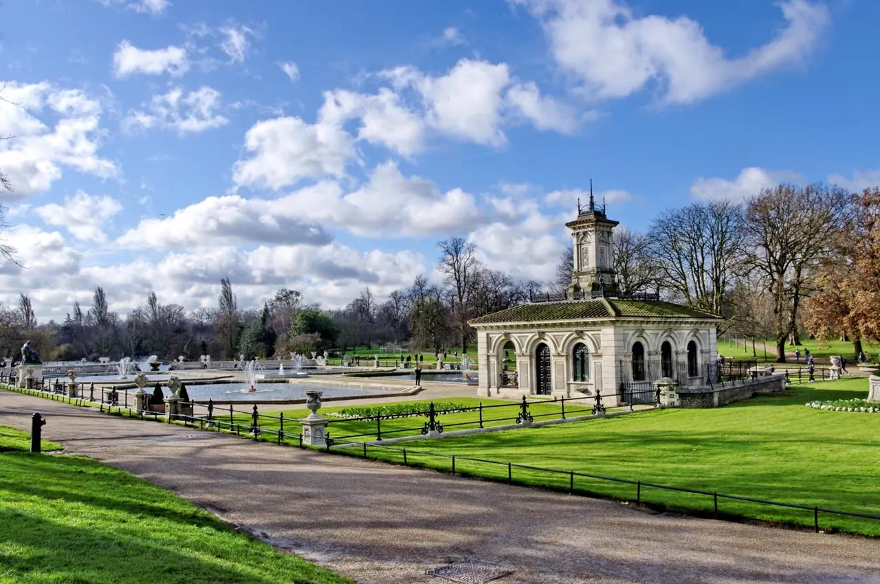 A formal garden with a fountain basin, a white ornate pavilion with a small tower, green lawns, iron railings and bare trees under a partly cloudy sky.