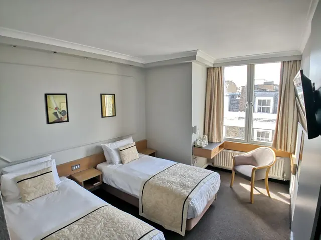 A twin bedroom with two single beds, a wall-mounted desk and armchair, a wall-mounted television, and a window looking out over neighbouring buildings.