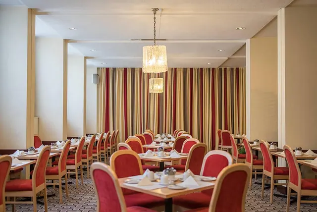 A dining room with red upholstered chairs, white-clothed tables set with napkins and cups, a crystal chandelier, and striped curtains on the back wall.