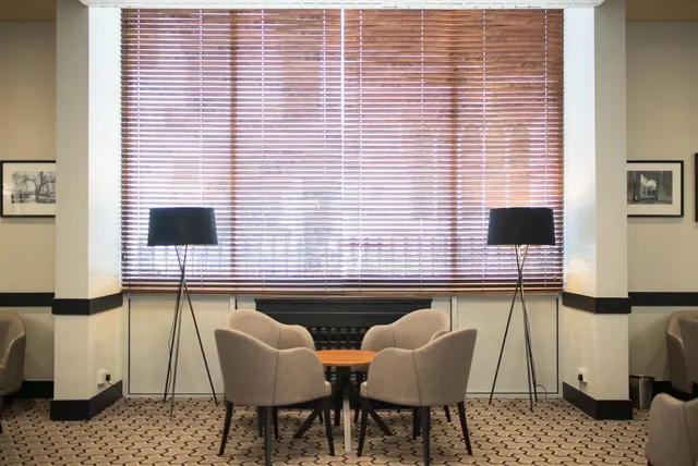 A lounge seating area with four grey armchairs around a small round wooden table, flanked by two floor lamps in front of a window with closed wooden blinds.