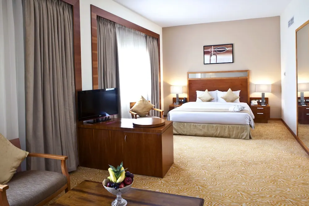 A hotel bedroom with a king bed, wooden headboard, bedside lamps, a television on a wooden cabinet, an armchair and a fruit bowl on a low wooden table.