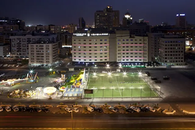 Night view from an upper floor across a floodlit football pitch and fairground rides toward the Landmark hotel building and the city skyline.