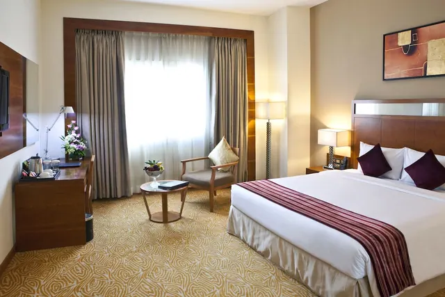 A staged hotel bedroom with a king bed, an armchair beside a round side table, a work desk with a kettle, and a window with sheer and dark curtains.