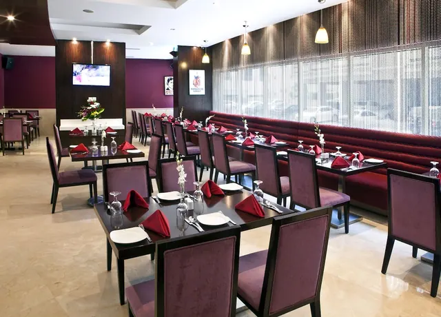 A restaurant dining room with set tables, red napkins, and a burgundy banquette along a wall of windows showing a street outside.
