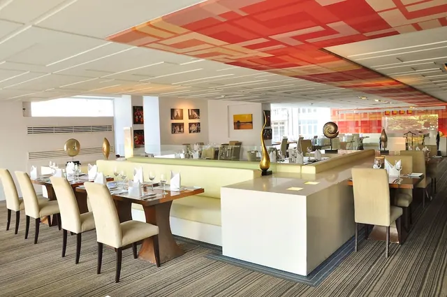 A restaurant dining room with set tables, upholstered chairs, a pale green banquette and a red patterned ceiling.