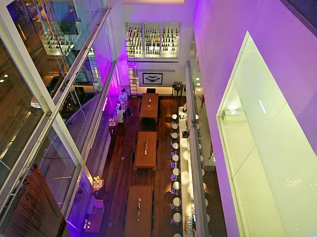 Aerial view down into a restaurant with long wooden tables, purple lighting, a wine wall and a row of bar stools along a counter.
