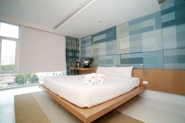 A white king bed with two towel animals sits on a raised wooden platform in a bedroom with a blue geometric wall panel, a desk, and a window showing trees and buildings outside.