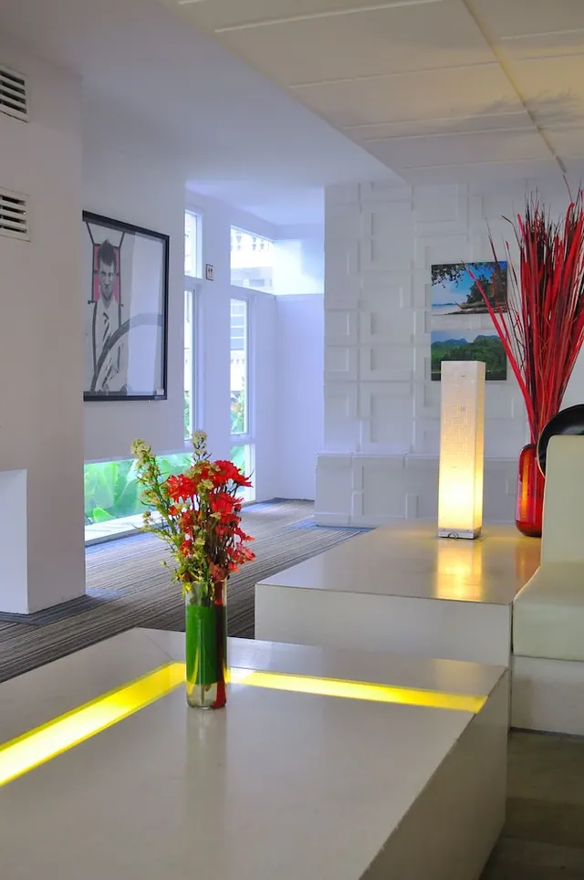 A white modern lobby seating area with illuminated low tables, a glowing floor lamp, a red flower arrangement, and windows showing green foliage outside.